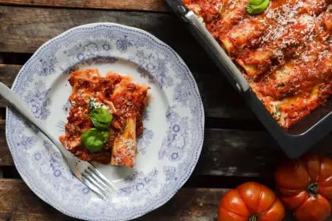 En portion läcker cannelloni med tomatsås och basilika på en vacker tallrik. Bredvid syns en ugnsform med mer cannelloni och färska tomater. Ett perfekt cannelloni recept!