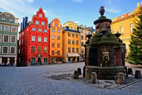 Färgglada hus i Gamla Stan, Stockholm, med en gammal fontän på kullerstensbelagd plats.