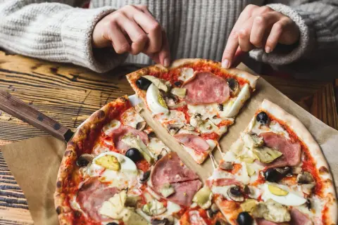 Une personne choisit une part de pizza avec une riche pizza garniture : jambon, œufs, olives, artichauts et champignons.