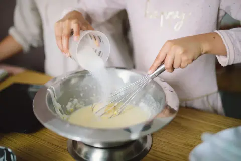Préparation de la pâte à crêpes : du sucre est versé dans un saladier où une main fouette la préparation.