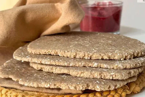 Una pila de pan sin levadura, rústico y con trozos de avena, junto a un vaso de mermelada de frutos rojos y una tela marrón.