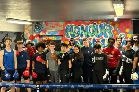 A group of young boxers, some wearing gloves, stand in a boxing ring. One boxer is in the foreground, ready to run.