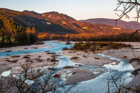 Le **saut de la Drôme** offre un paysage de rivière aux eaux bleues vives serpentant entre des bancs de galets, sous des montagnes dorées par le soleil couchant.