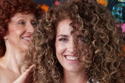 Dos mujeres sonríen, una con cabello rizado pelirrojo y la otra con cabello rizado castaño, mostrando estilos de cortes de pelo afro.