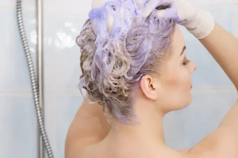 Kobieta w rękawiczkach nakłada fioletowy szampon na blond włosy. Czy szampon koloryzujący niszczy włosy?