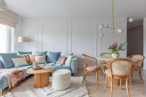 Miękka, błękitna sofa i pudroworóżowe akcenty tworzą przytulną atmosferę. Ciepłe kolory do salonu podkreślają drewniane meble i jasny dywan.