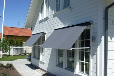 Grå markiser monterade på ett vitt trähus ger skugga åt fönstren.
