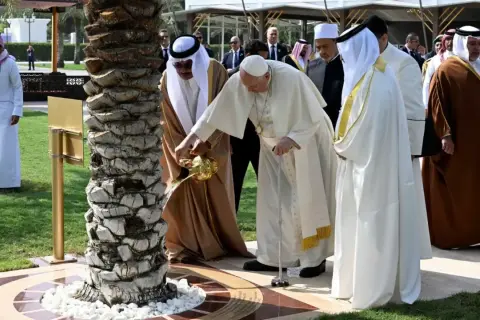 Papież Franciszek i przywódca Zjednoczonych Emiratów Arabskich wspólnie podlewają drzewo. W tle widać ludzi w tradycyjnych strojach, jakby na spotkaniu z muezinem.