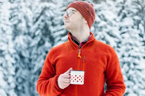 Mężczyzna w pomarańczowym polarze i czapce trzyma kubek w śnieżnym lesie. Polar jest wykonany z miękkiego, ciepłego materiału.