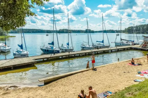 Letnie popołudnie na **jeziorach na Mazurach**. Drewniane molo, zacumowane żaglówki i plażowicze cieszący się słońcem.