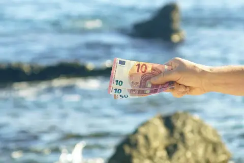 Dłoń trzyma banknoty euro na tle morza. W Czarnogórze obowiązuje euro, więc te pieniądze przydadzą się na wakacjach.