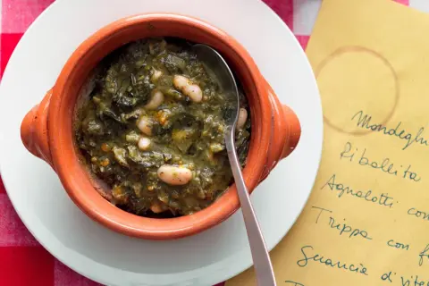 Zuppa di cavolo nero e fagioli in ciotola di terracotta, servita su un piatto bianco con tovaglia a quadri rossi. Accanto, un foglio giallo con un elenco di piatti.