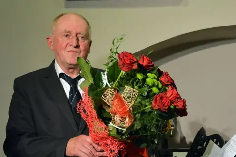 Starszy mężczyzna w garniturze trzyma bukiet czerwonych róż i zielonych kwiatów.