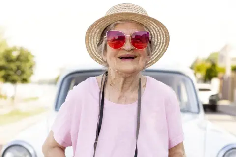 Une femme souriante, chapeau de paille et lunettes roses, prête pour partir seule en vacances à 60 ans. Une voiture ancienne en arrière-plan.