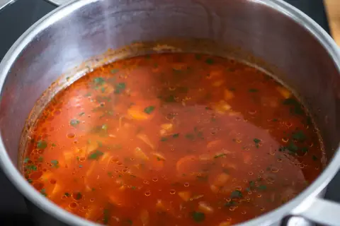 Gorąca zupa pomidorowa w garnku, z widocznymi kawałkami warzyw i zieleniną. Czy zupę pomidorową zaprawia się mąką?