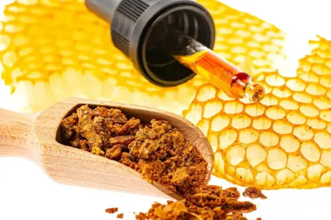 Propolis co to? Złota kropla z pipety na plastrze miodu, obok drewnianej łyżki z pyłkiem.