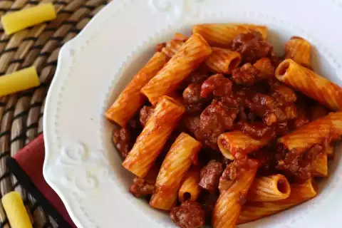 Piatto di pasta con salsiccia e sugo rosso, servita in un piatto bianco decorato.