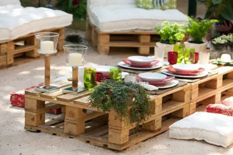 Mesa de jardín con ideas con palets, decorada con vajilla, velas y plantas. Cojines a los lados invitan a relajarse.