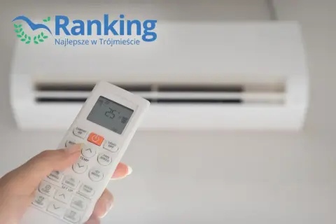 Dłoń trzymająca pilot do klimatyzacji przed białym urządzeniem.
