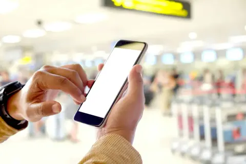 Ręce trzymają smartfon z pustym ekranem. W tle widać zamazany terminal lotniczy, sugerujący, że to idealny moment na **lot odprawa online kiedy** tylko chcesz.