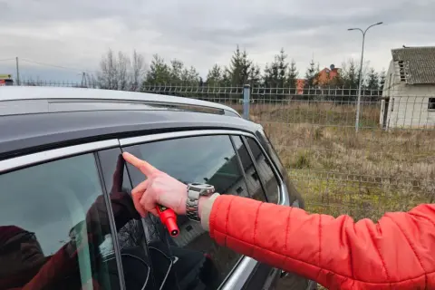 Mężczyzna w czerwonej kurtce wskazuje palcem na drzwi swojego pierwszego samochodu, trzymając w dłoni czerwony przyrząd.