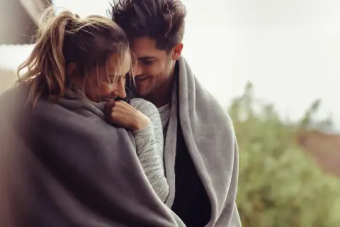 Coppia abbracciata sotto una coperta, sorrisi complici e sguardi teneri, un dolce inizio di una relazione.