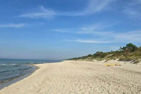 Słoneczny dzień nad zalewem kamieńskim. Szeroka, piaszczysta plaża z łagodnymi falami i zielonymi wydmami w tle. Kilka osób odpoczywa na piasku.