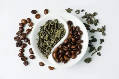 En vit skål delad i två, med grönt te och kaffebönor. En evig kamp mellan te eller kaffe.