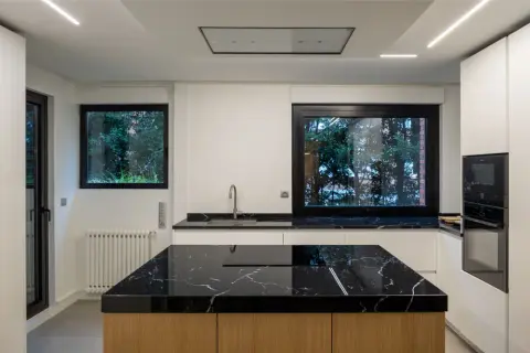 Moderna cocina con isla central de mármol negro y madera. Encimeras a juego, electrodomésticos integrados y grandes ventanales.