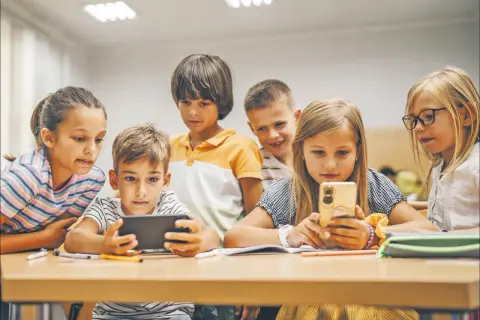 Dzieci skupione na ekranach telefonów i tabletu, uczące się i bawiące razem. Wspólna **socjalizacja** w cyfrowym świecie.