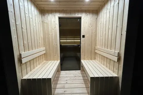 Drewniana sauna z ławami i szklanymi drzwiami. Dyskretna wentylacja w saunie zapewnia komfort.