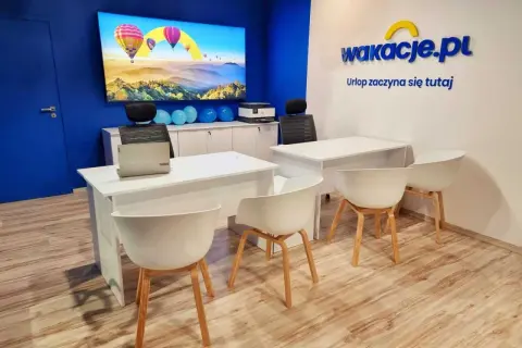 Nowoczesne biuro z logo "wakacje.pl", ekranem z balonami i białymi biurkami.