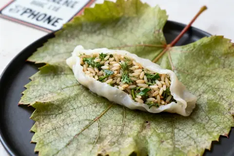 Une feuille de vigne farcie, garnie de riz et d'herbes fraîches, repose sur une grande feuille de vigne verte sur une assiette noire.