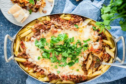 En stor ugnsform med gyllene penne, täckt med köttfärssås och smält ost. En portion pasta al forno är upplagd på en tallrik bredvid.