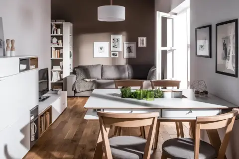 Nowoczesny, prostokątny salon z jadalnią. Szara kanapa, białe meble, drewniana podłoga i jasne dodatki tworzą przytulną atmosferę.