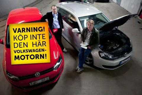 Två män står vid bilar. En röd bil har en gul skylt med texten "VARNING! KÖP INTE DEN HÄR VOLKSWAGEN-MOTORN!". En silverfärgad bil har motorhuven öppen.