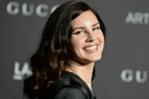 Uśmiechnięta Lana Del Rey, artystka znana z nastrojowych piosenek, na tle logo Gucci.