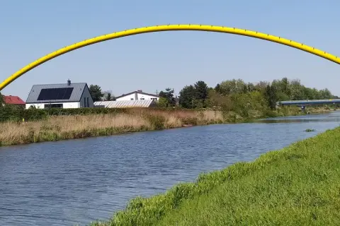 Żółty łuk mostu nad kanałem żerańskim, z domami i zielenią w tle.