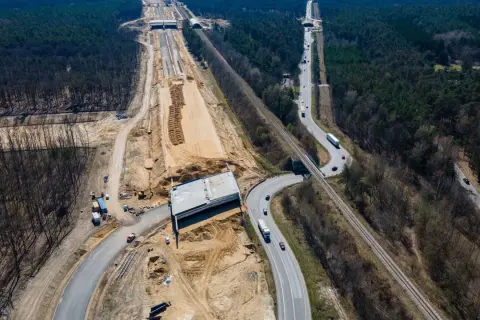 Budowa drogi ekspresowej S19 Via Carpatia w lesie. Widoczna nowa nawierzchnia, most, kolej i ruch samochodowy.