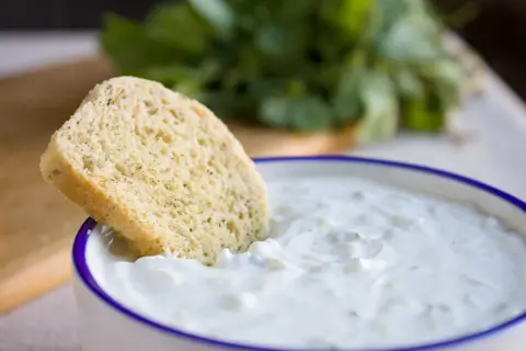 Ein Stück Brot wird in eine Schüssel mit cremigem, selbstgemachtem Zaziki getunkt. Frische Kräuter im Hintergrund.