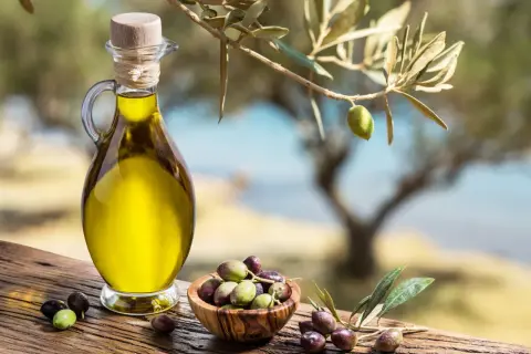Caraffa di **olio di oliva** dorato accanto a una ciotola di olive fresche, sotto un ramo d'ulivo.