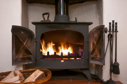 Ein Holzofen mit brennendem Feuer. Daneben ein Korb mit **Holz** und ein Werkzeugset. Die **Asche** glüht im Feuer.