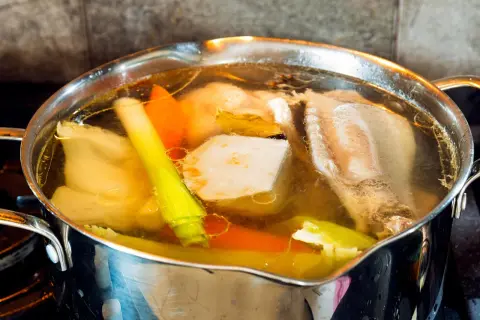 Mętny rosół bulgocze w garnku, unosząc aromat warzyw i mięsa. Widać kawałki marchewki, pora i kości.