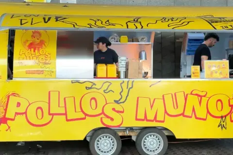 Camión amarillo "Pollos Muñoz" con un gallo rojo. Dos empleados trabajan en el interior.