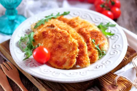 Delizioso pollo impanato al forno, croccante e dorato, servito con pomodorini e rucola su un piatto bianco.