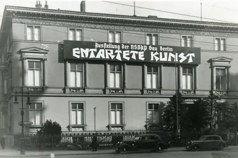 Große Banner verkünden die "Ausstellung der NSDAP Gau Berlin ENTTARTETE KUNST". Autos und Fahrräder stehen davor.