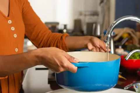 Une personne remplit une cocotte bleue sous un robinet pour faire bouillir de l'eau chaude.