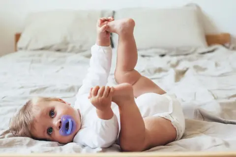 En 11 månaders bebis med napp ligger på rygg på en säng och leker med sina fötter.