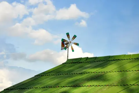 Piccolo impianto eolico domestico su tetto verde, con pale colorate che girano nel cielo azzurro con nuvole bianche.