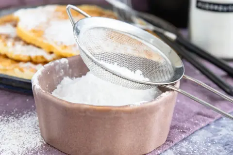 Feiner Puderzucker rieselt aus einem Sieb in eine Schale. So einfach lässt sich puderzucker selber machen für leckere Waffeln.